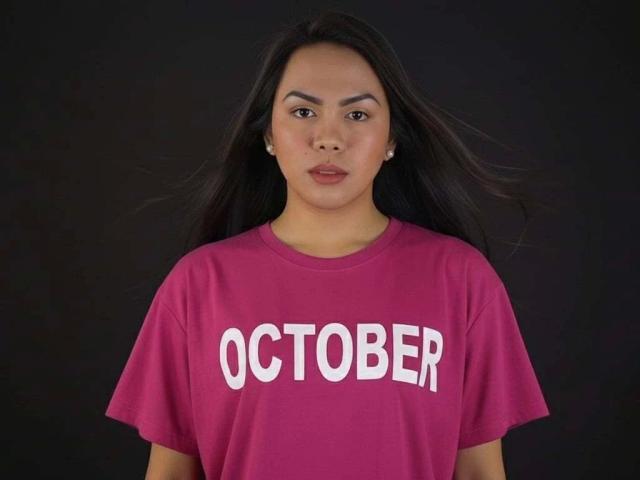 AbbyOBierna-hot has long black hair, brown eyes, and wears a pink shirt with white text. She has a neutral expression and small pearl earrings.
