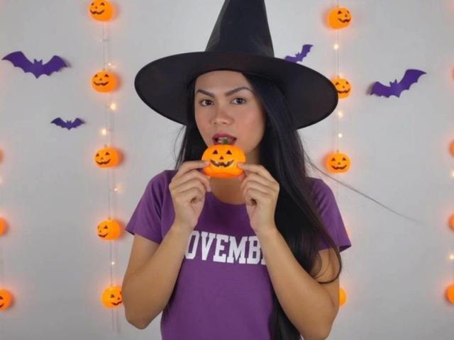 AbbyOBierna-hot with long black hair, wearing a black witch hat and purple shirt, eating a small orange pumpkin-shaped candy, with purple bat decorations and string lights in background.