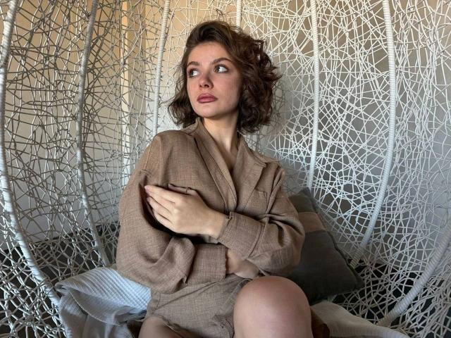 IceFoxy-hot with wavy brown hair, wearing a brown, oversized blazer, sitting in a white, woven hanging chair, covering her small tits with one hand, looking to the side.