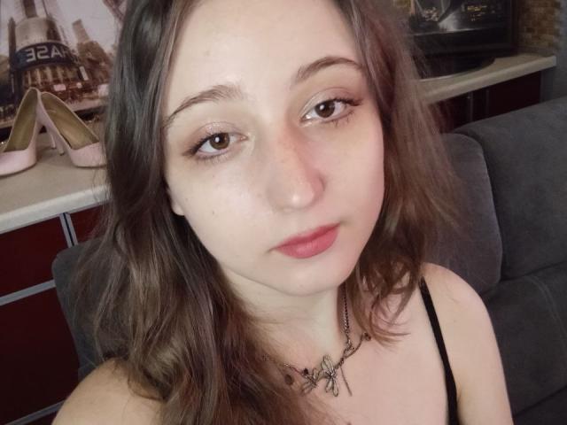 TwinkyKiss has pale skin, brown eyes, and wavy brown hair. She's wearing a black spaghetti strap top with a necklace, and her pink lips are slightly parted.