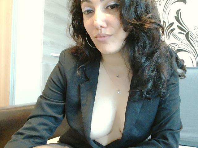 SexyMirra, avec ses cheveux noirs bouclés, porte un blazer noir, laissant entrevoir ses seins volumineux et un petit piercing au mamelon. Elle a des boucles d'oreilles cercles et un sourire discret..