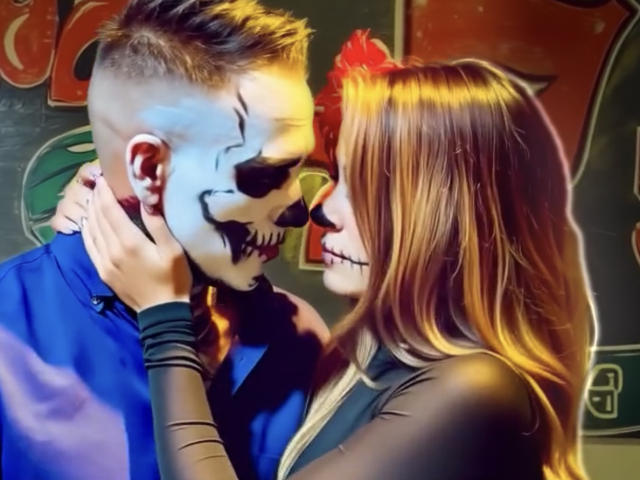 BigJLila with dirty blonde hair, in a black top, holds a guy's face painted as a creepy clown with white makeup and black nose, in a blue shirt.