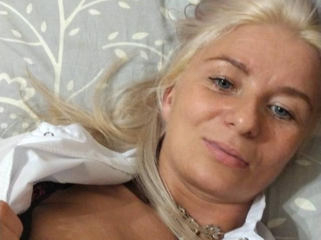 RoseeZ has blonde hair, blue eyes, and a slight smile, lying down with a white shirt partially open, showing a bit of her chest and a necklace.
