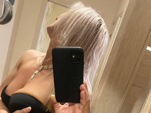 RoseeZ taking a mirror selfie, showing off her big tits in a black strapless top, with long blonde hair covering her face, holding a black phone with a textured case.