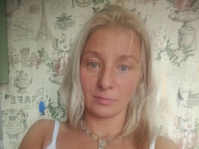 RoseeZ has blonde hair, blue eyes, and a slight smile. She's wearing a white tank top with a silver necklace, and her tits are small. She looks cute.