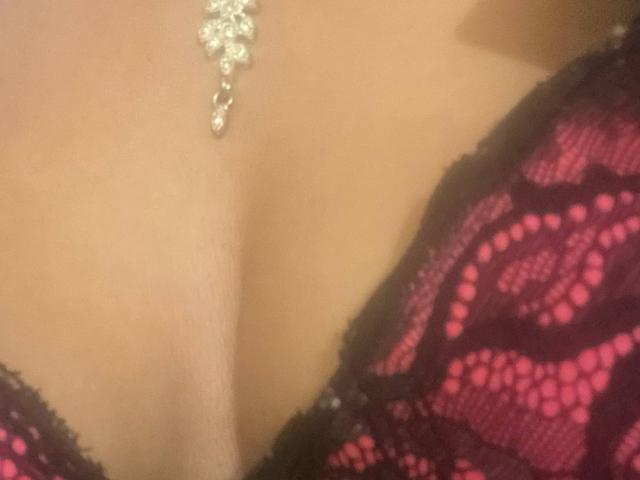 RoseeZ's tits in a red lace bra with black trim, showing a delicate silver cross necklace hanging between her cleavage. Soft, warm skin.