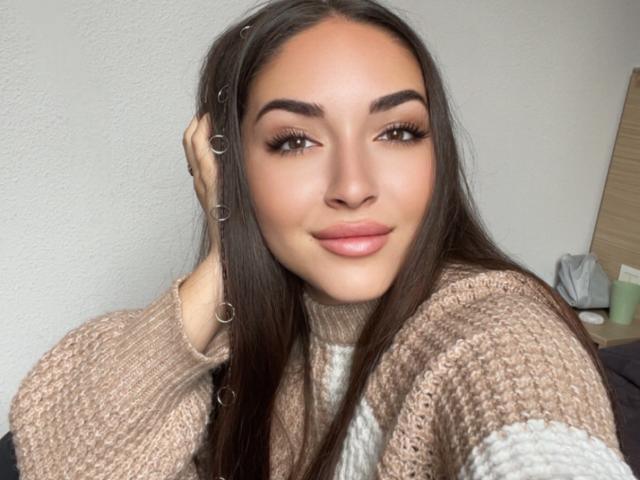 AquaLady has long brown hair, brown eyes, and full pink lips, wearing a beige knit sweater with metal hairpins. She's smiling softly, looking at the camera.