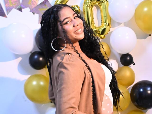 SofiFoxy69 with long black curly hair, big hoop earrings, smiling, wearing a brown cardigan over a white dress, surrounded by yellow, black, and white balloons.