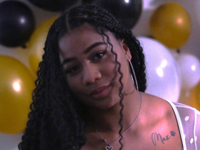 SofiFoxy69 has dark curly hair, wearing a white top, with a tattoo on her chest, and large hoop earrings, looking down with a slight smile. Balloons in the background.