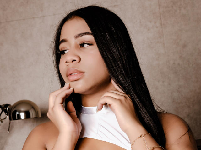 SofiFoxy69 with long black hair, light brown skin, and full lips, touching her chin with both hands, wearing a white halter top, showing her small tits, and a gold bracelet.