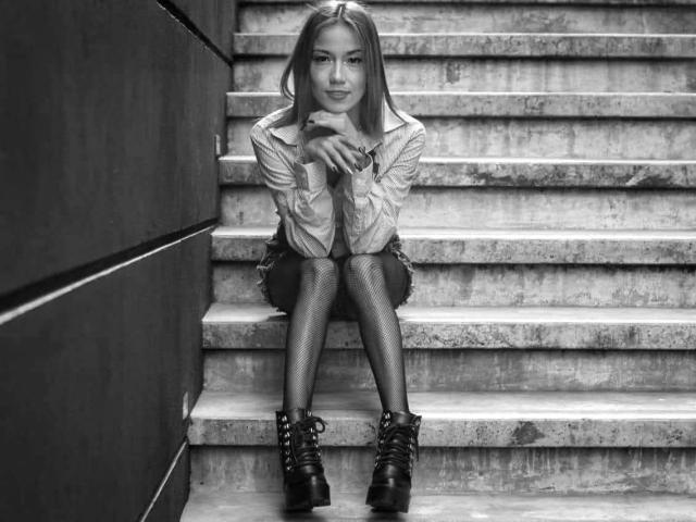 Veroo69 with long hair, wearing an open shirt, fishnet stockings, and black lace-up boots, sits on concrete stairs, hands near face, smiling subtly, in a black-and-white photo.