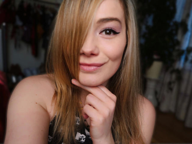 Noukonekox-hot with blonde hair, pink eyeshadow, and a cute smile, her hand on her chin, showing off her soft pink lips and slight nose freckle.