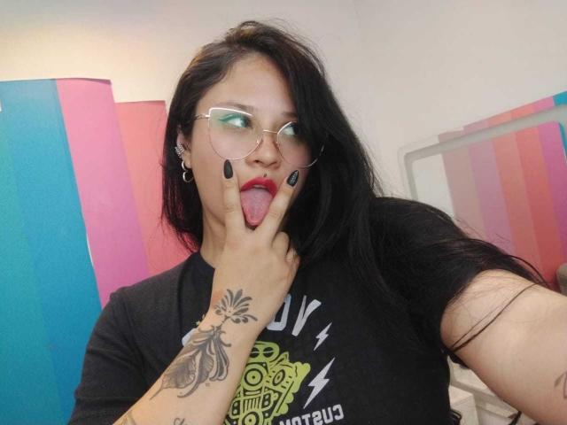 MaraVixen with black hair, glasses, and red lipstick, sticking out tongue, making "V" sign with black-painted nails, showing tattoo on hand, wearing black shirt.