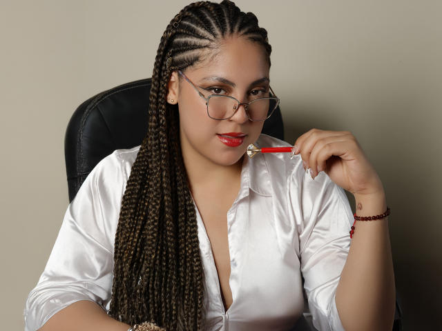VictoriaKurt with long braids, wearing glasses and a white shirt, biting a red pen, her red lips and bracelet visible, sitting in a black chair.