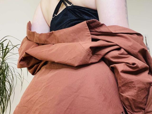 KarKutie with pale skin, black top, and a burnt-orange dress, arms crossed, showing her back and a tattoo on her right arm, looking down at the camera.