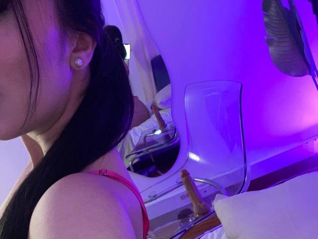 Antonella Watson, com cabelo preto preso em um rabo de cavalo, vestindo um sutiã vermelho, mostrando o ombro nu e um brinco de diamante, refletida em um espelho com uma cadeira transparente..