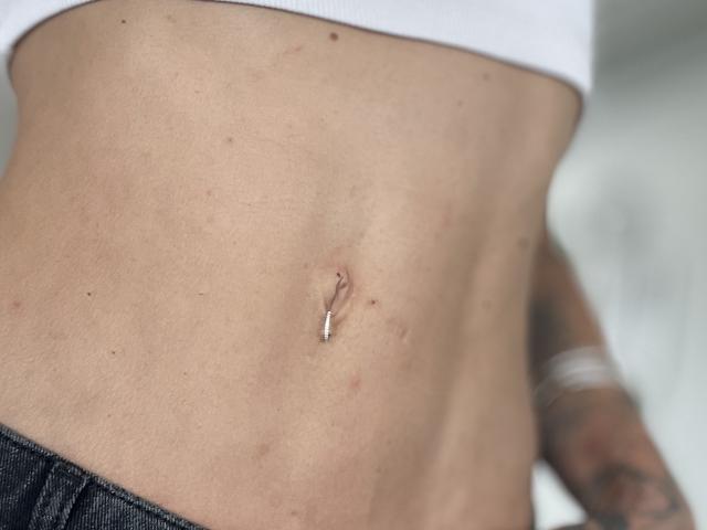 La pancia piatta e leggermente abbronzata di DaneeSoHot, con un piccolo piercing all'ombelico lucido e una maglietta bianca, indossa pantaloni neri, mostrando un braccio tatuato sul lato destro..