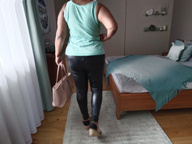 NaturalWoman with a curvy, muscular butt wearing a green tank top and black leather pants, holding a pink bag, standing on a wooden floor in a bedroom.