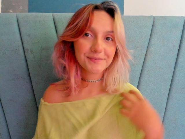 AlterEgoSecond has pink and blonde hair, a cute smile, and a choker. She's wearing a yellow off-shoulder top, showing a small amount of her tits.