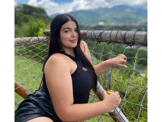 LaraJames with long black hair, wearing a black sleeveless dress, sitting on a wooden fence with a wire mesh, holding it with her right hand, smiling.