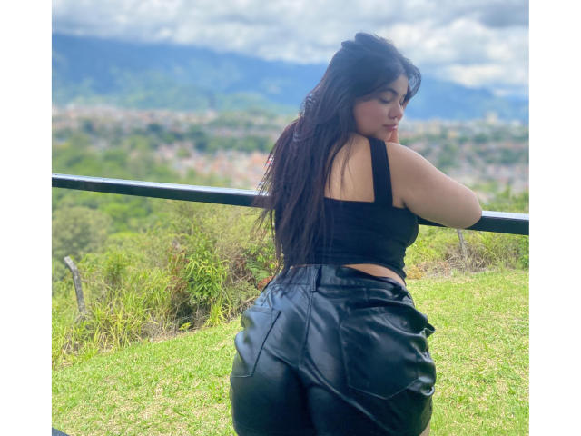 LaraJames with long dark hair, black sleeveless top, and black leather pants, showing her ass, leaning on a rail, looking over her shoulder, outdoors, mountains in background.