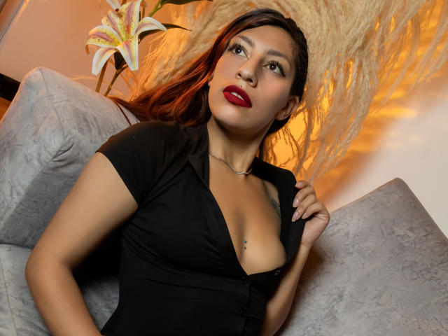 AlisonCold with red lipstick, black button-up dress, and cleavage, sitting on a grey couch, hand on chest, lily in background, warm light, brown hair, looking up.