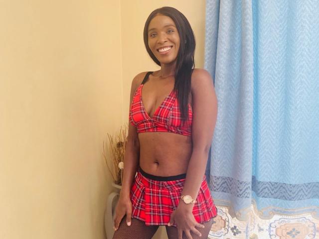 BrookeCherryX has dark skin, long black hair, and is smiling while wearing a red plaid bra and skirt, showing her tits and small ass. She's wearing a watch.