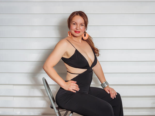 VictoriiaJones with red hair, black cut-out dress, showing her tits, sitting on a chair, wearing large orange earrings, and black pants, smiling at the camera.