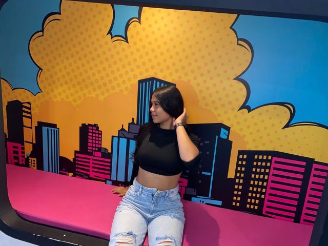 GenesisIsis, com cabelo preto longo, top preto e calças jeans rasgadas, sentada em uma superfície rosa, olhando para o lado, com a mão no cabelo, em um mural colorido da cidade, em estilo pop art..