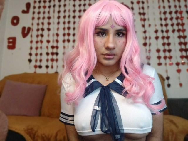 MelinoHottie with pink wig, wearing a white crop top with navy blue ribbon, showing her tits, standing in front of a brown couch, with heart decorations in the background.