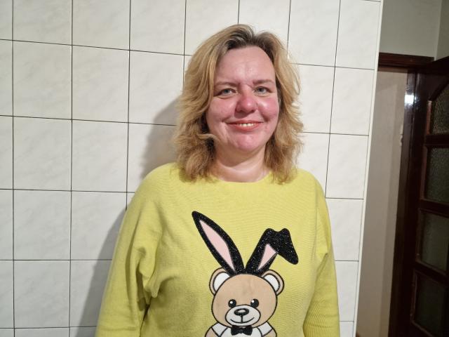 VestaWoody with blonde, wavy hair, smiling, wearing a yellow sweatshirt with a black and pink bunny graphic, standing against white tiled wall.
