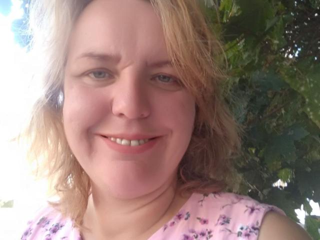 VestaWoody has light blonde, wavy hair, blue eyes, and a pink floral shirt, smiling with a slight squint, showing small, white teeth and a cute, chubby face.