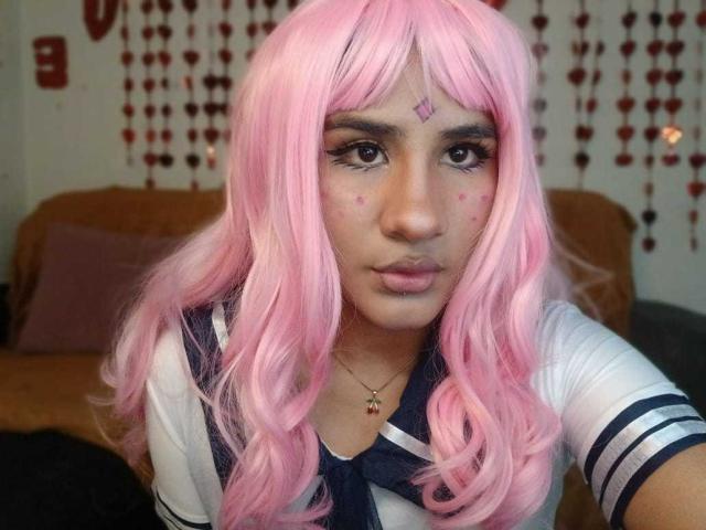 MelinoHottie with pink wig, white sailor top, and makeup dots on face, looking at camera with pouty lips, necklace with red and gold beads, brown couch in background.