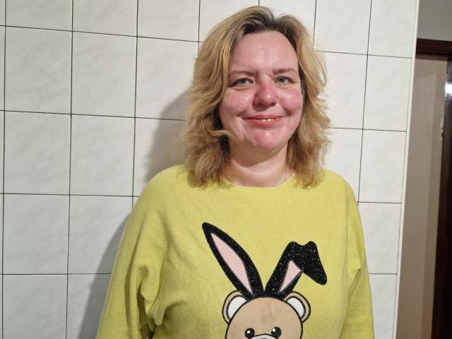 VestaWoody with blonde hair, smiling, wearing a yellow sweatshirt with a black bunny face, standing against a white tiled wall. She has pink cheeks and a cute, relaxed look.
