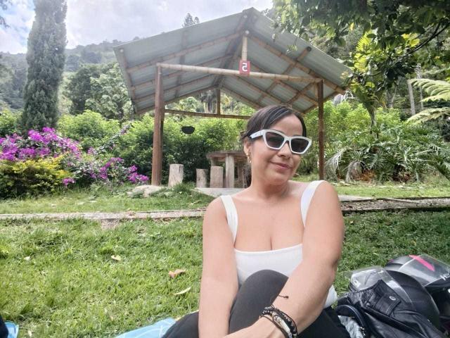 TamaraFoxy with white sunglasses, white tank top showing her boobs, black pants, sitting on grass, green background, wooden structure, backpack on right, smiling, dark hair in a bun.