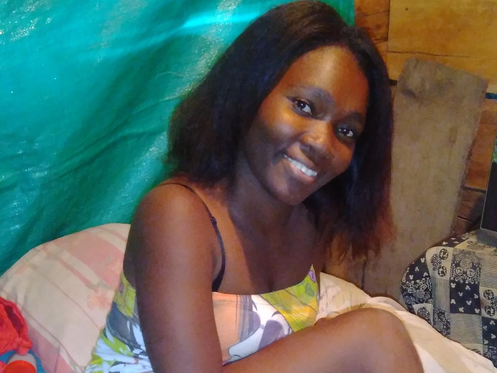 BriyiX has dark brown skin, medium-length black hair, and is smiling with white teeth. She's wearing a colorful dress, showing her small tits and black bra straps.