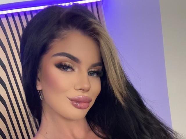 IvyRaven has long black and blonde hair, glossy pink lips, and heavy eyeliner. Her tits are subtle but noticeable. She's looking at the camera with a slight smile.