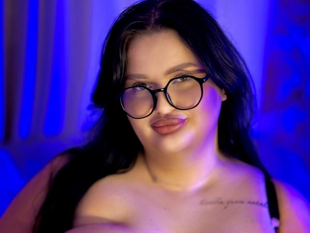 MonaSteele with black hair, wearing glasses, pouty lips, and a black strap top, showing off her tits, tattoo on her chest, in blue and purple lighting.