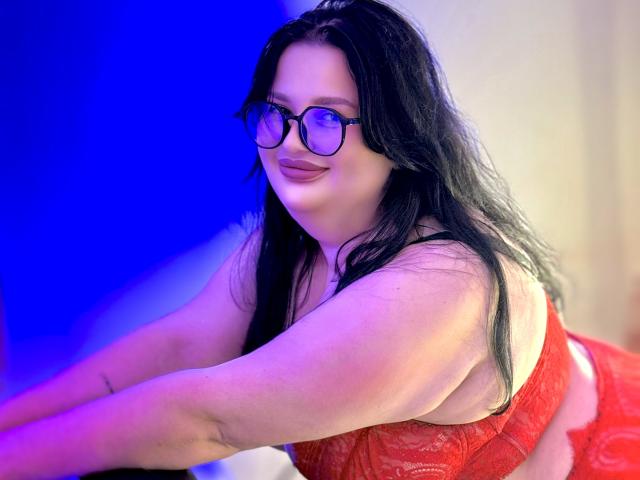 MonaSteele with black hair, glasses, and red lace lingerie, leaning forward, showing her curvy body and large tits. Blue light in background. Smiling, confident.