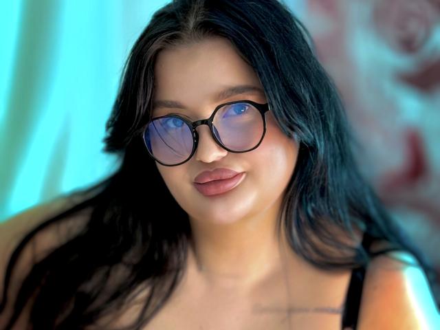 MonaSteele with black hair, blue-tinted glasses, and pouty lips, wearing a black top, showing a subtle tattoo on her chest. Her tits are small and her pussy is not visible.