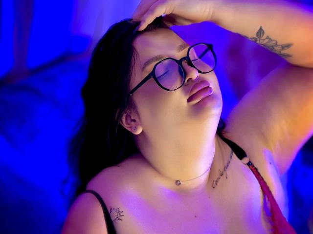 MonaSteele with glasses, black hair, and tattoos, laying back with eyes closed, head tilted, hand on forehead, blue and purple lighting, wearing red lace bra, slightly open mouth.