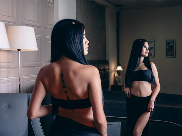 Danielle Harper, avec ses longs cheveux noirs, porte un haut noir sans manches qui dévoile sa poitrine, une jupe noire fendue sur la cuisse, et un tatouage noir sur le dos. Elle se tient devant un miroir..