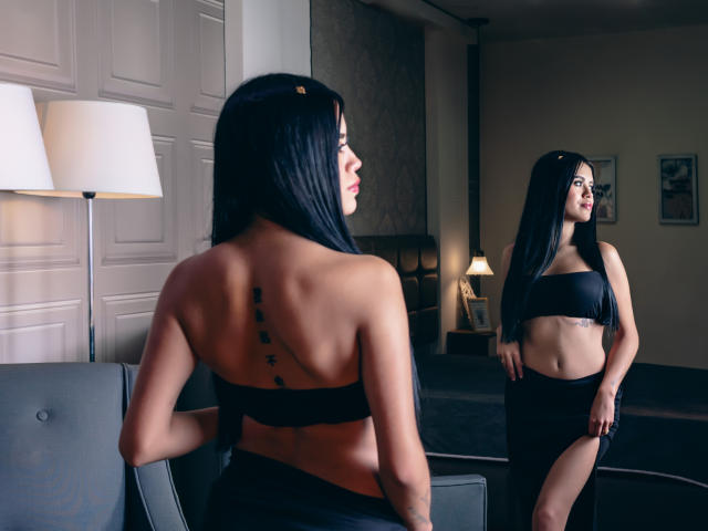 DanielleHarper with long black hair, back tattoo, black strapless top, black skirt, standing in front of mirror, showing her toned ass and flat stomach, in dimly lit room.