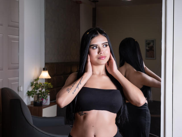 DanielleHarper with long black hair, wearing a black strapless top, hands on her face, reflection in mirror, tattoo on right arm, subtle smile, in a softly lit room.