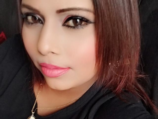 SexyInaBebe has pink lipstick, black eyeliner, and brown eyes with straight brown hair, wearing a black shirt, and a gold necklace with a small cross.