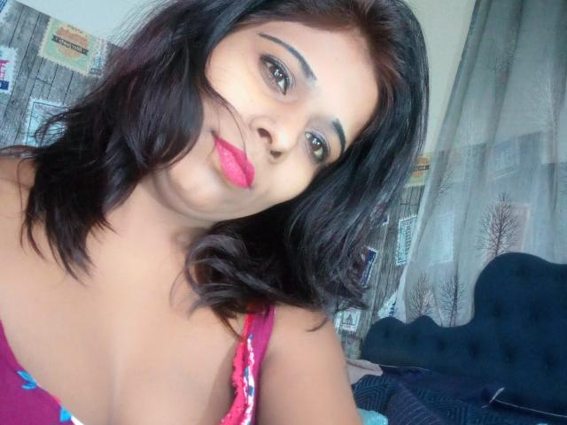SexyInaBebe with dark brown hair, pink lipstick, and thick eyebrows, wearing a pink floral top, showing a hint of her small tits, in a bedroom.