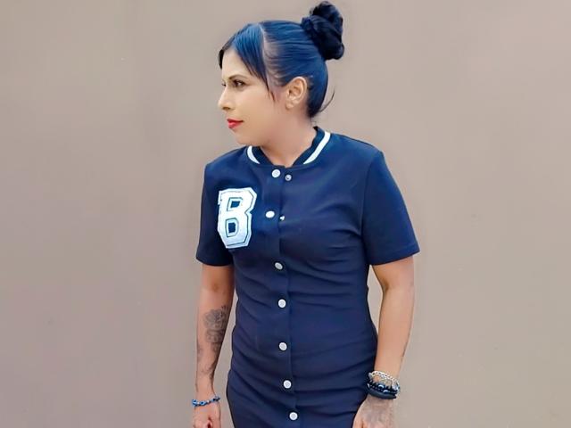 SexyInaBebe with dark blue sports dress, white buttons, and "B" patch, black bun, tattooed arm, red lipstick, and bracelets, standing against plain background.