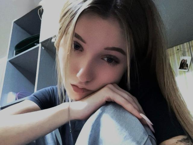 SilverxKitty with long blonde hair, looking down, resting her chin on her hand, wearing a black shirt, grey pants, and a thin bracelet, in a slightly dim room.