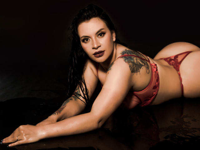 KatalinaJohnson with wet black hair, lying on her stomach in red lace lingerie, showing her tattoos, butt, and looking at the camera with dark lipstick.