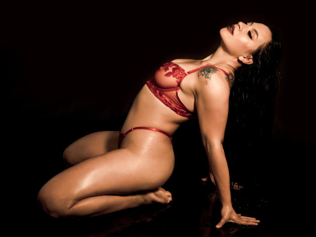 KatalinaJohnson kneels in red lace lingerie, back arched, eyes closed, wet skin glistening, tattoo on left shoulder, hands on black surface, head tilted up, looking sexy.